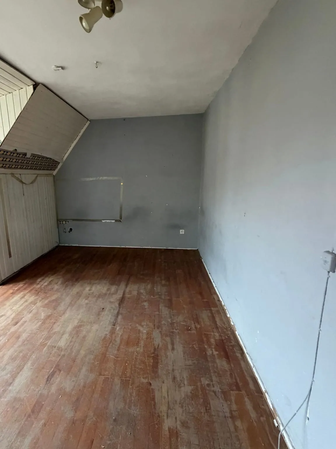 Dasselbe Dachzimmer leer geräumt, mit freiem Holzboden, graublauen Wänden und weiß verkleideter Dachschräge.