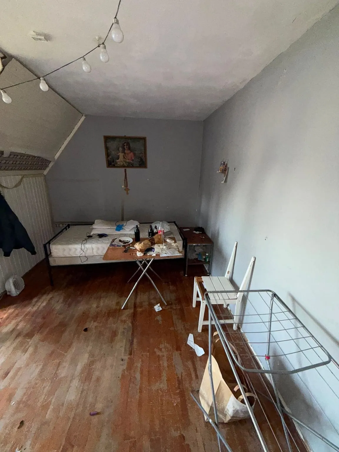 Dachzimmer mit Bett, Klapptisch mit Flaschen und Geschirr, Nachttisch, weißem Stuhl und Wäscheständer, an der Rückwand ein gerahmtes Bild und an der Decke eine Lichterkette.