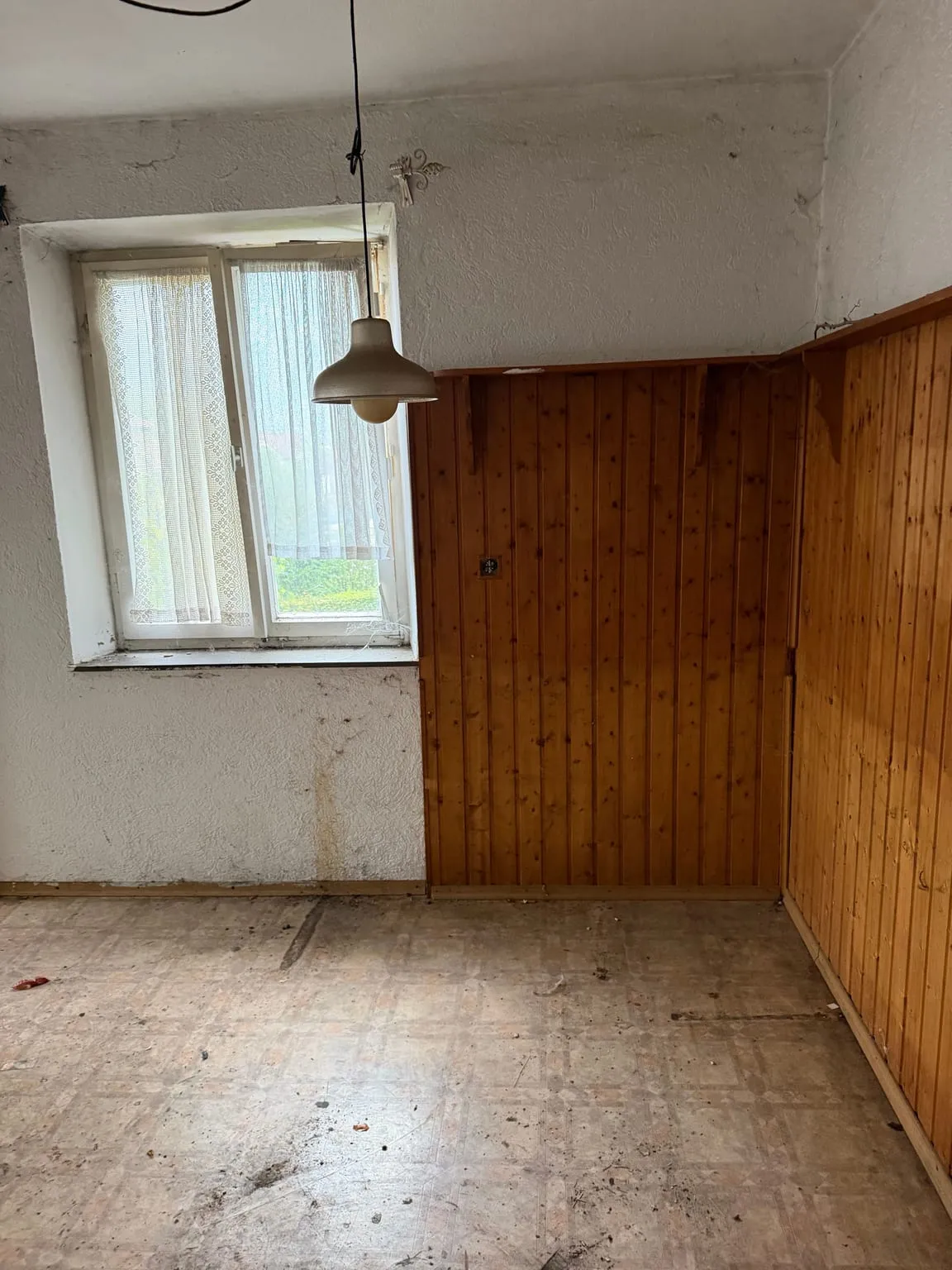 Leere Essecke mit Holzvertäfelung über Eck, Fenster mit Spitzengardine, Pendelleuchte und freiem Boden.