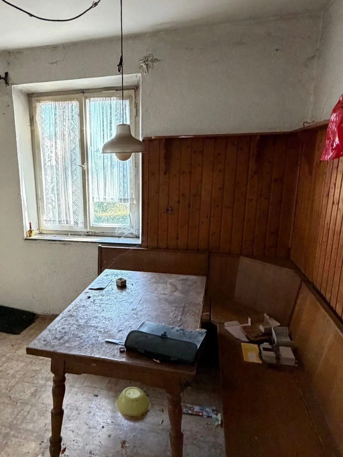 Essecke mit Holztisch, Eckbank vor einer Holzvertäfelung, Kartons und Plastiktüten auf Bank und Boden, dahinter ein Fenster mit Spitzengardine.