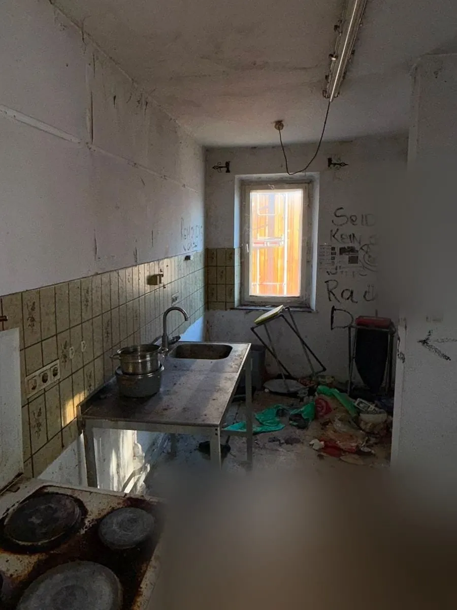Schmale Küche mit verschmutztem Elektroherd, Spültisch aus Edelstahl, umgekipptem Gestell und Unrat auf dem Boden vor dem Fenster.