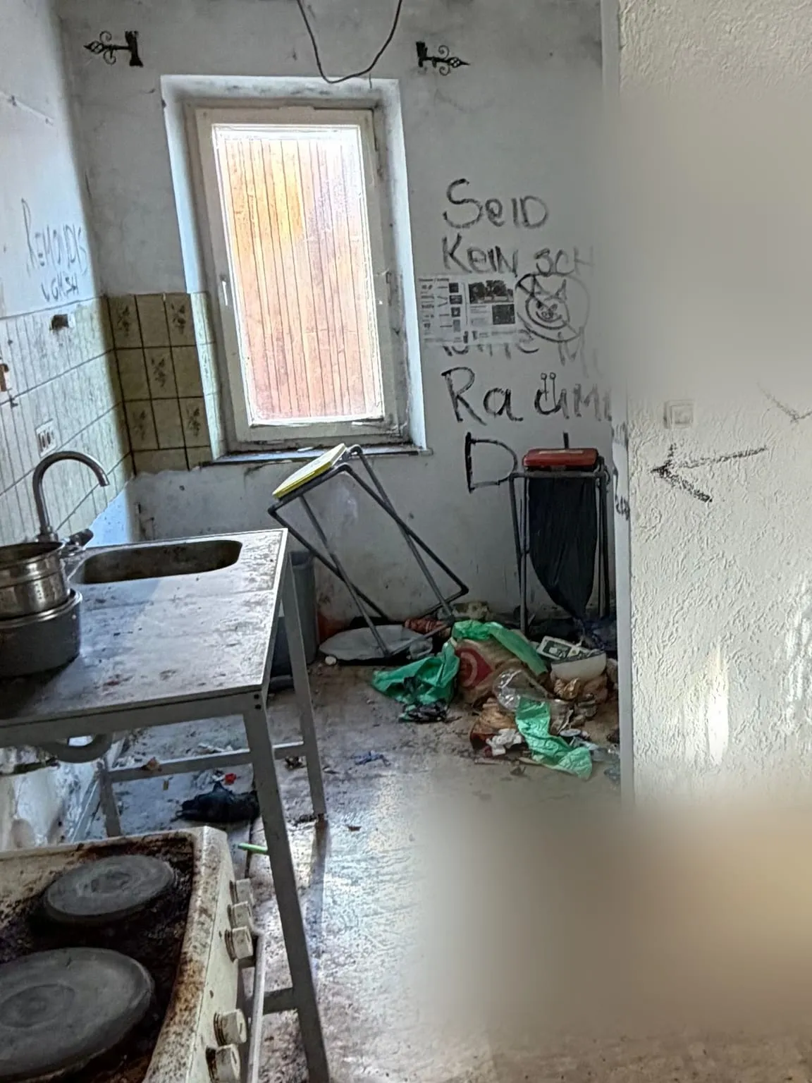 Küche vor der Räumung: Unter dem Fenster stehen eine Spüle mit Töpfen, ein Herd, ein Gestell und ein Abfalleimer, am Boden liegen Tüten und Abfall.
