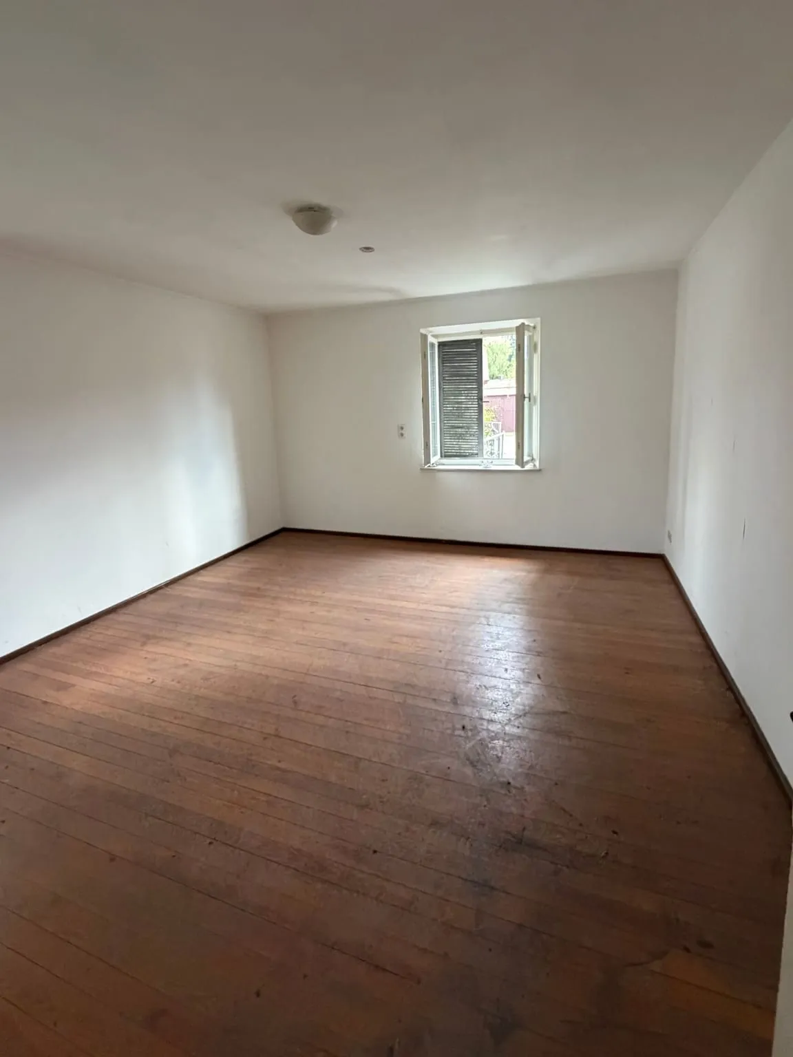 Dasselbe Mehrbettzimmer nach der Räumung, leer mit Dielenboden, weißen Wänden und geöffnetem Fenster.
