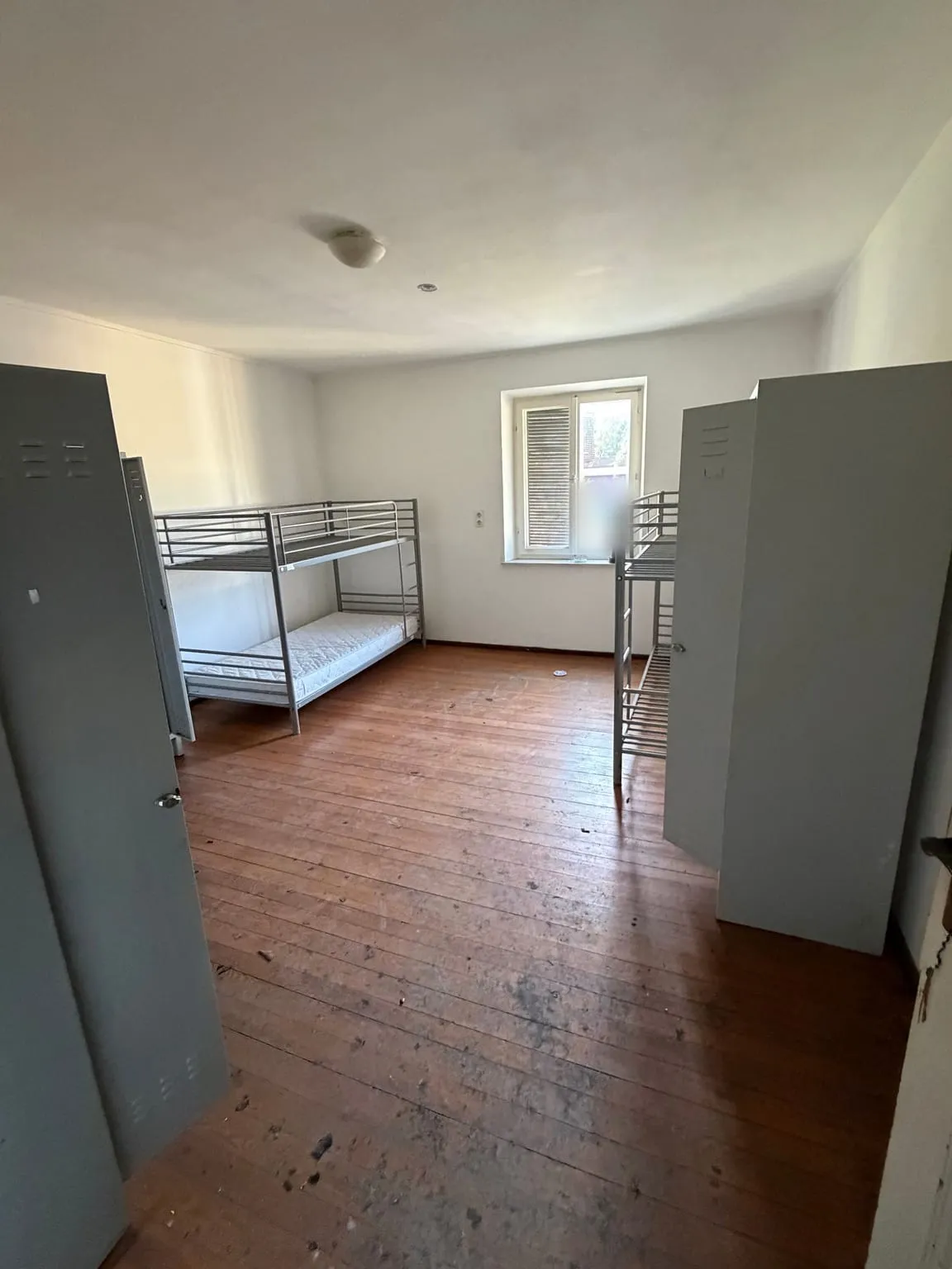 Mehrbettzimmer mit Dielenboden, zwei Doppelstockbetten aus Metall und mehreren grauen Spinden vor einem Fenster mit Klappladen.