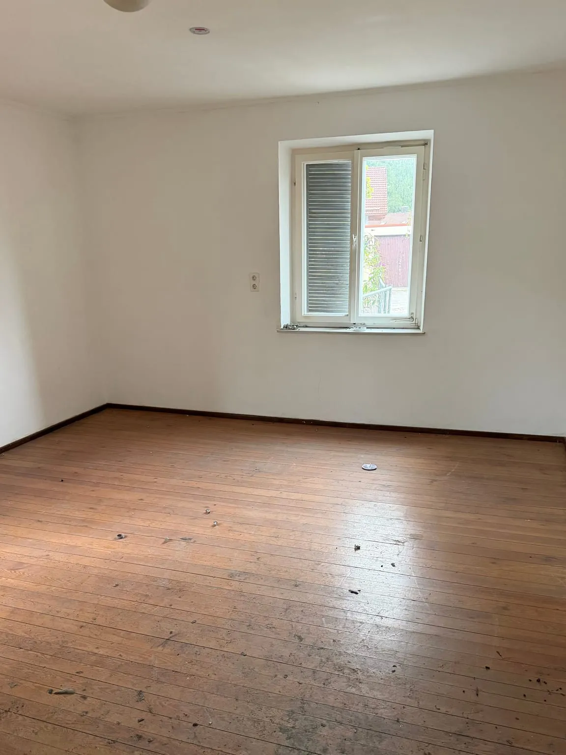 Dasselbe Zimmer vollständig leer geräumt, mit freiem Dielenboden und weißen Wänden unter dem Fenster mit Rollladen.