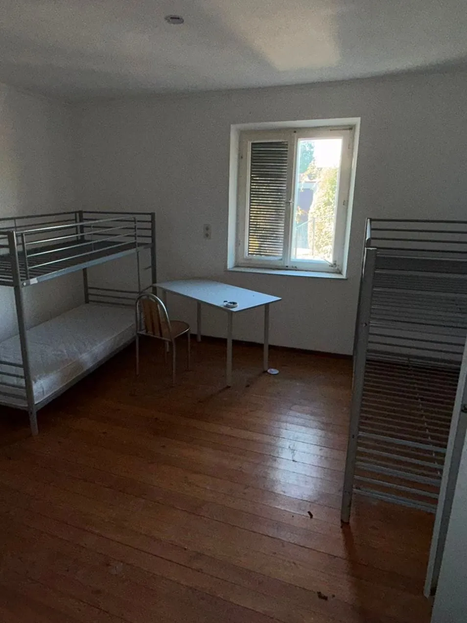 Zimmer mit zwei Etagenbetten aus Metall, einem weißen Tisch und einem Stuhl vor dem Fenster sowie einem Spind am rechten Bildrand.