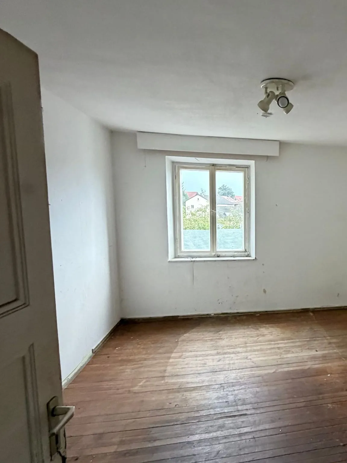 Dasselbe Zimmer leer geräumt, mit freiem Holzdielenboden, weissen Wänden und dem Fenster unter dem Rollladenkasten.