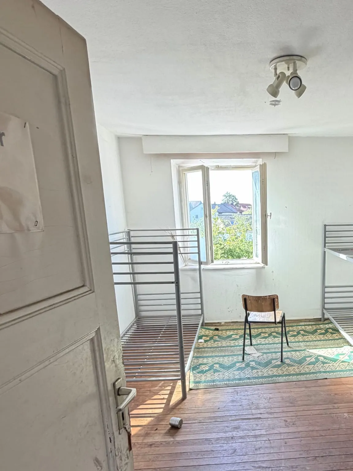 Zimmer mit zwei Etagenbetten aus Metall, einem Holzstuhl auf einem grün gemusterten Teppich und dem Dielenboden, links die geöffnete Zimmertür.