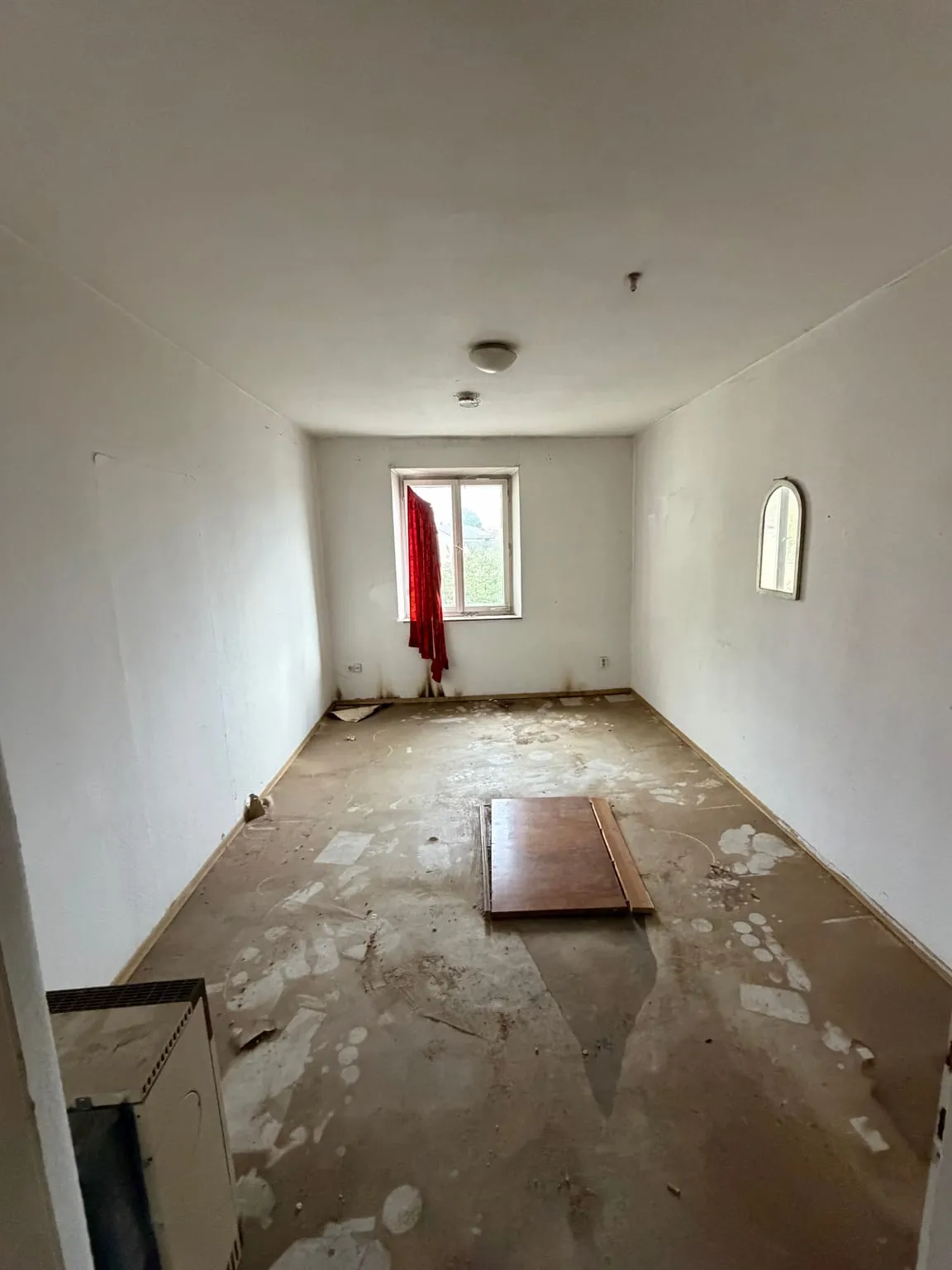 Dasselbe Zimmer leer geräumt, mit freiem Boden, einem Bogenspiegel an der rechten Wand und dem Fenster mit rotem Vorhang an der Stirnseite.