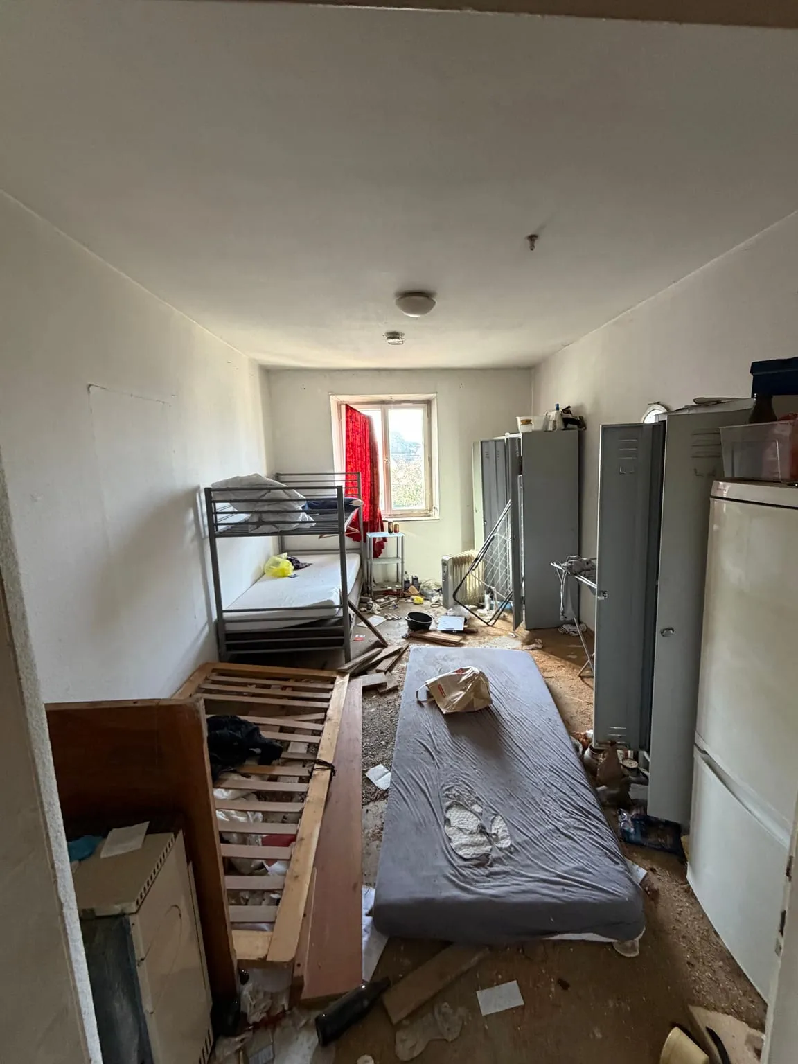 Schmales Zimmer mit Etagenbett, Metallspinden, Kühlschrank, einer Matratze und einem Lattenrost auf dem verschmutzten Boden, hinten ein Fenster mit rotem Vorhang.