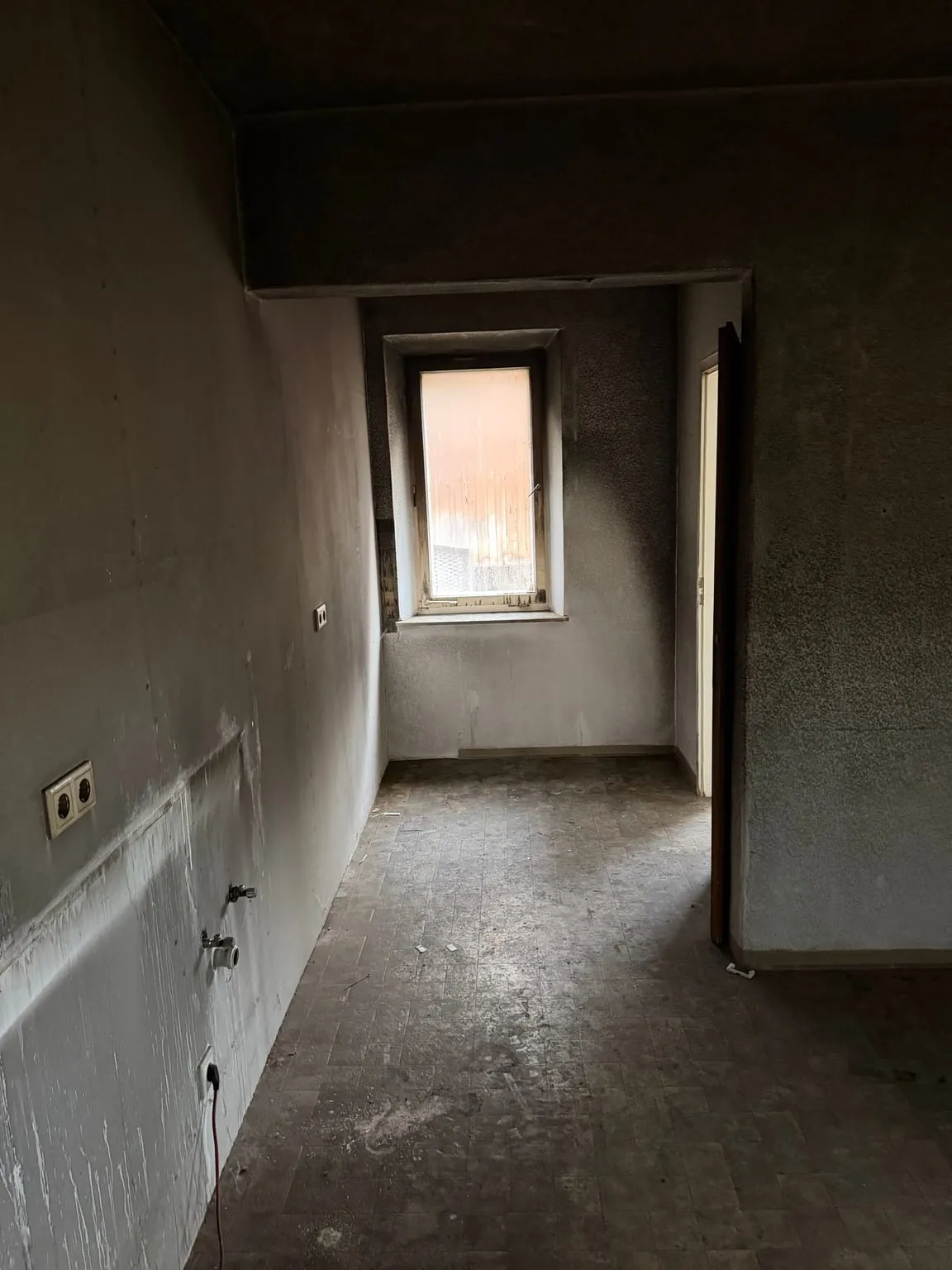 Dieselbe Küche leer geräumt, mit Strom- und Wasseranschlüssen an der linken Wand und freiem Blick durch den Durchgang zum Fenster der Nische.