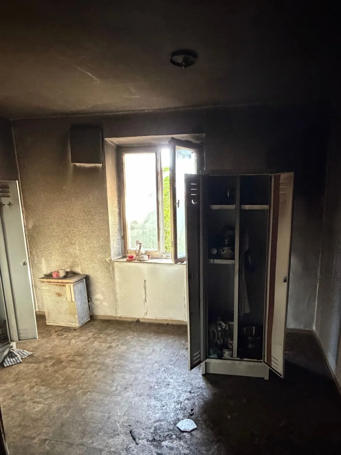 Verrußtes Zimmer mit zwei geöffneten Metallspinden, einem kleinen Gitterkasten unter dem Fenster und Resten auf dem Fliesenboden.
