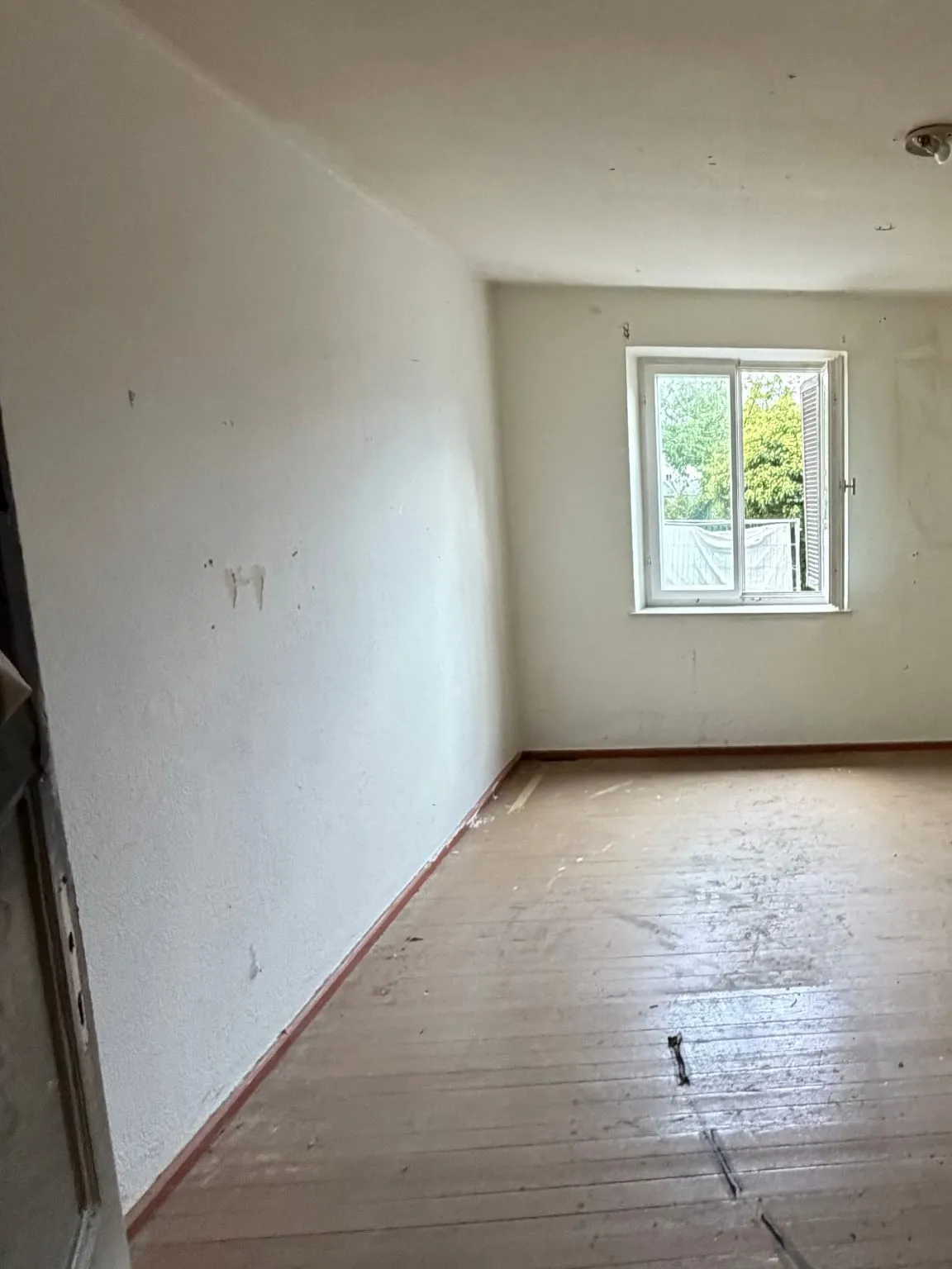 Dasselbe Zimmer nach der Räumung: leere weiße Wand, freier Dielenboden und das Fenster mit geöffnetem Flügel
