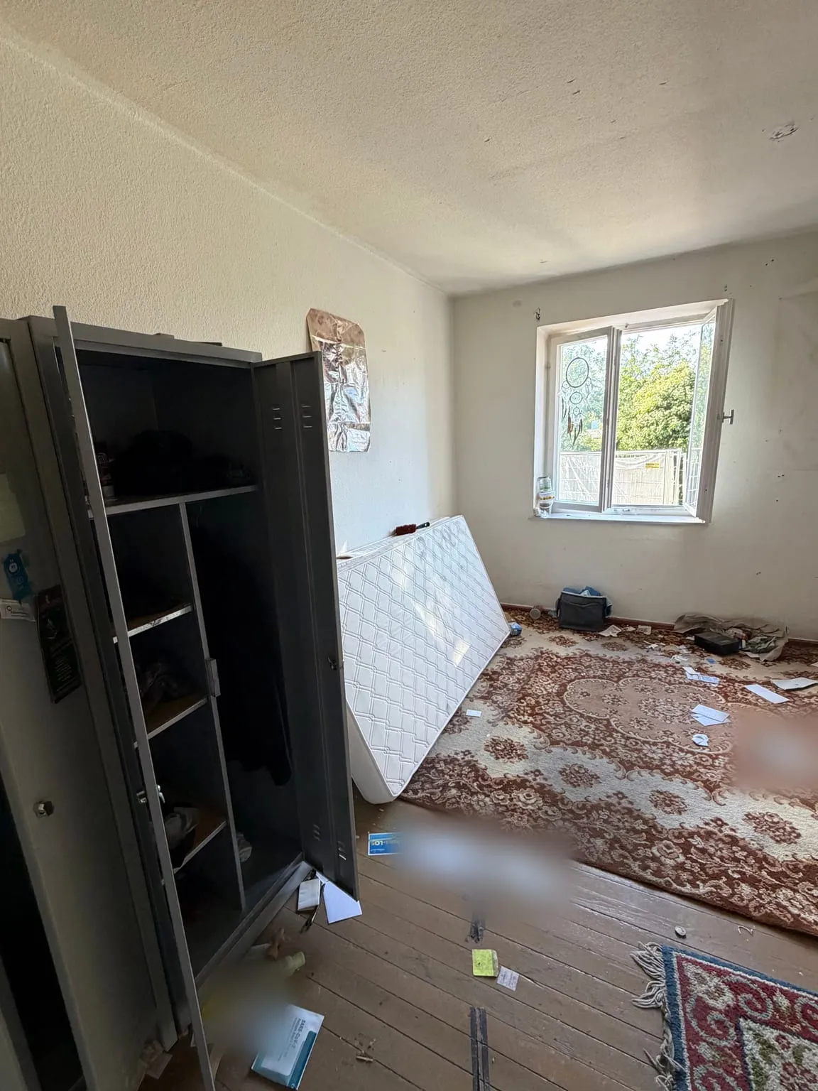 Zimmer vor der Räumung: geöffnete Metallspinde, eine an die Wand gelehnte Matratze, Teppiche und verstreute Papiere auf dem Holzboden, im Hintergrund ein Fenster