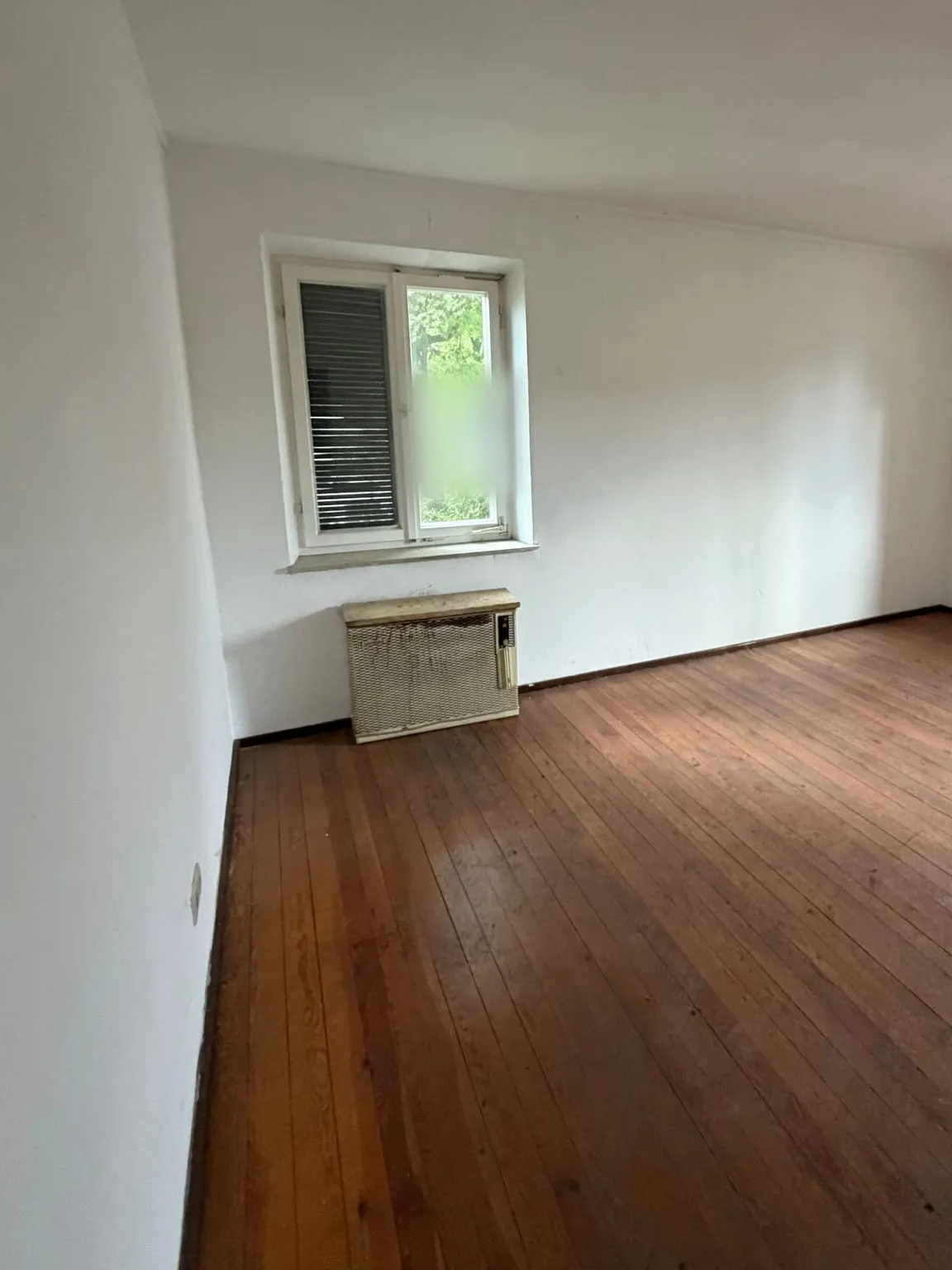 Dasselbe Zimmer leer geräumt, mit freiem Dielenboden und dem Heizgerät unter dem Fenster.