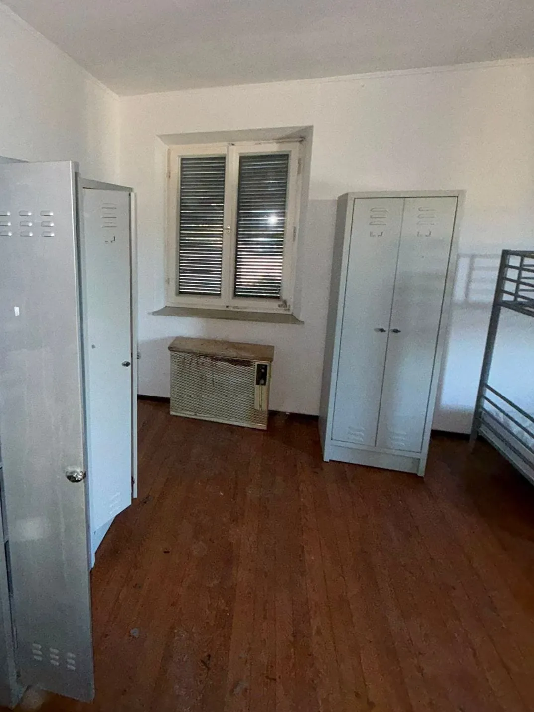 Zimmer mit Dielenboden, geöffneten Metallspinden links, einem Doppelspind rechts neben dem Fenster und einem Etagenbett am rechten Rand.