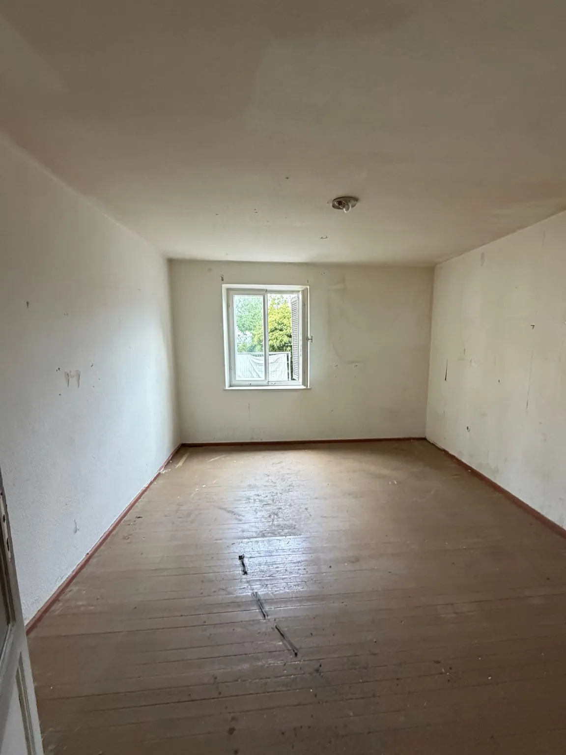 Dasselbe Zimmer vollständig leer geräumt, mit freiem Holzdielenboden, kahlen hellen Wänden und dem Fenster an der Stirnwand.