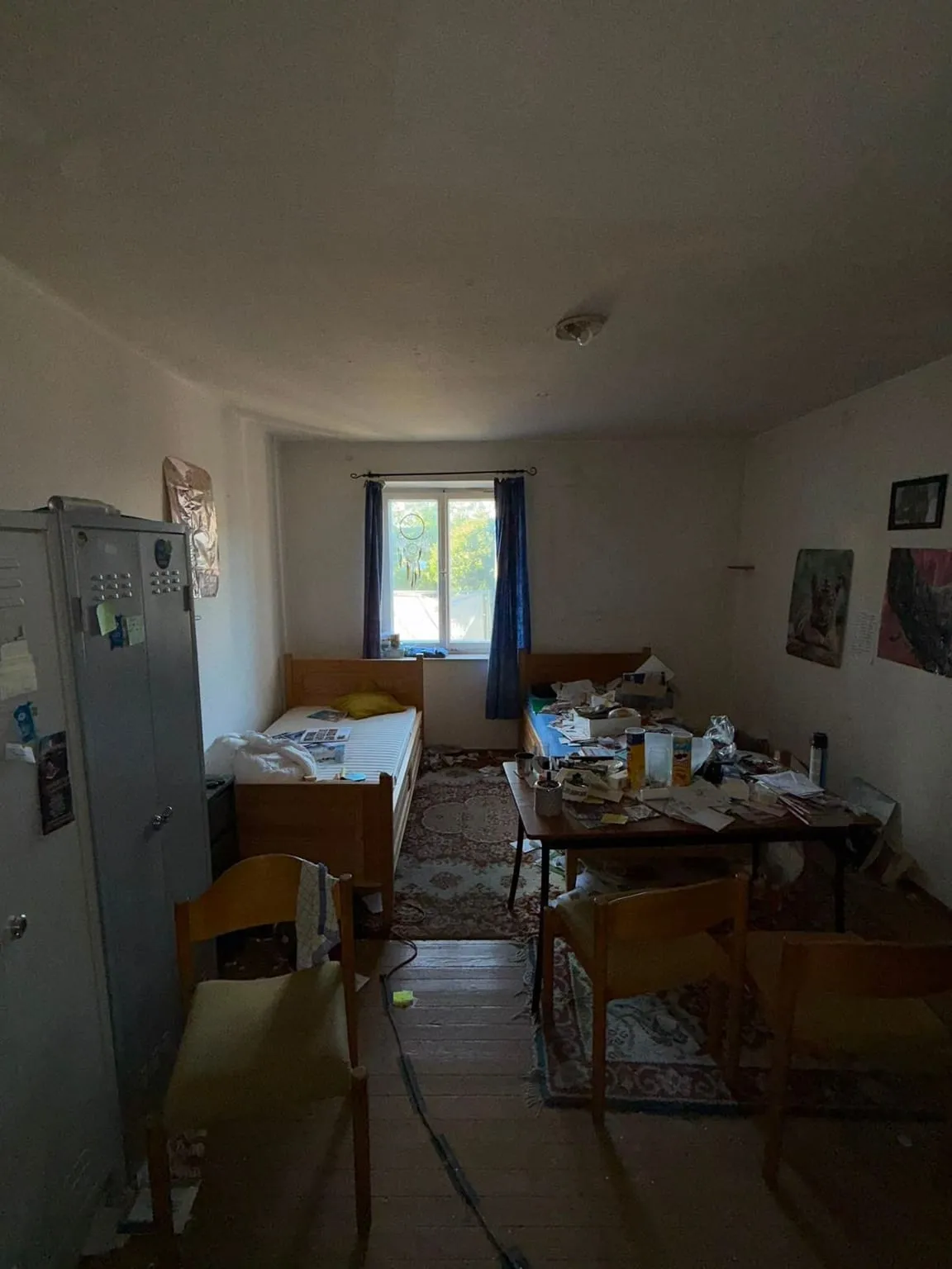 Längliches Zimmer mit Fenster an der Stirnwand, darin zwei Holzbetten, ein grauer Metallspind, ein Tisch mit Papieren und Flaschen, Holzstühle und Teppiche auf dem Dielenboden.