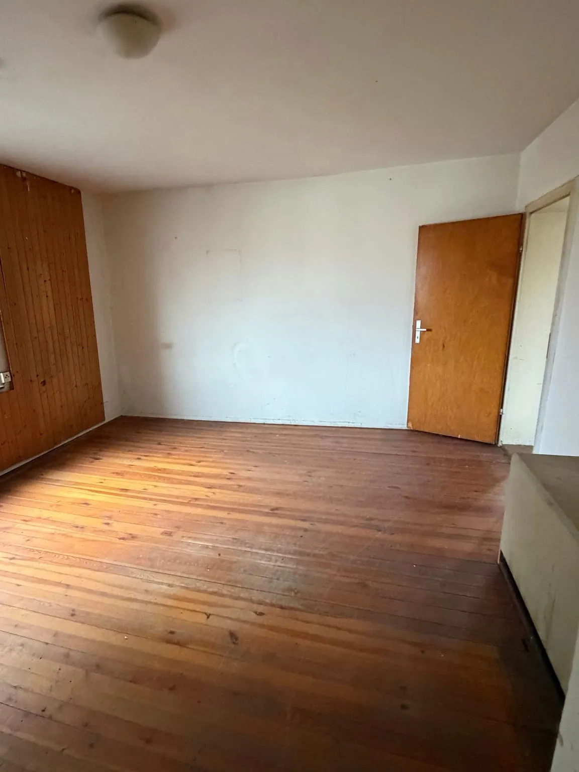 Dasselbe Zimmer leer geräumt, mit freien Holzdielen, Holzverkleidung an der linken Wand und geschlossener Holztür hinten rechts.