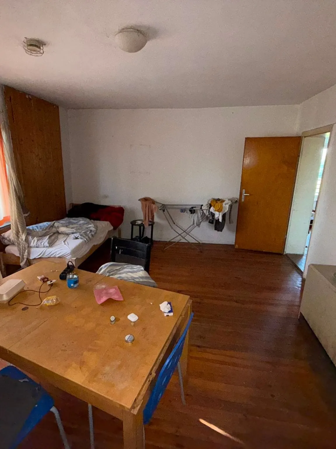 Zimmer mit Holzdielen, Esstisch mit Kleinteilen und blauen Stühlen im Vordergrund, dahinter ein Bett, ein Wäscheständer mit Kleidung und eine geschlossene Holztür.