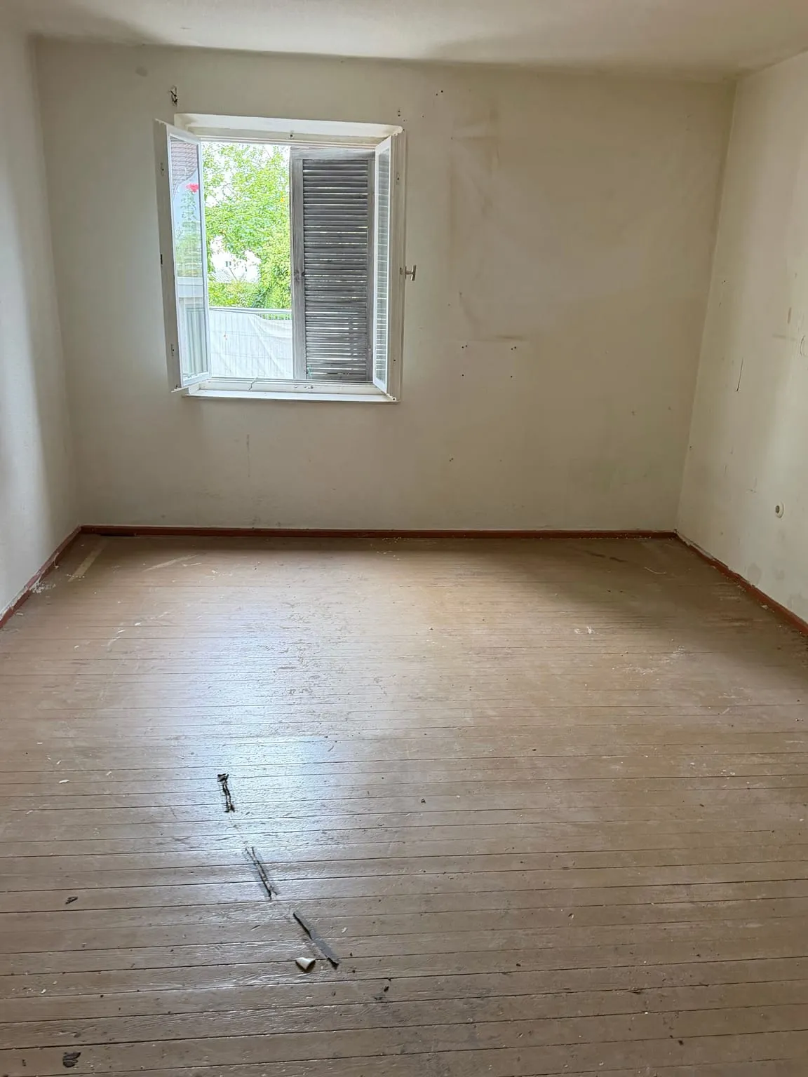 Dasselbe Zimmer leer geräumt, mit freiem grauem Holzdielenboden und kahlen Wänden unter dem geöffneten Fenster.