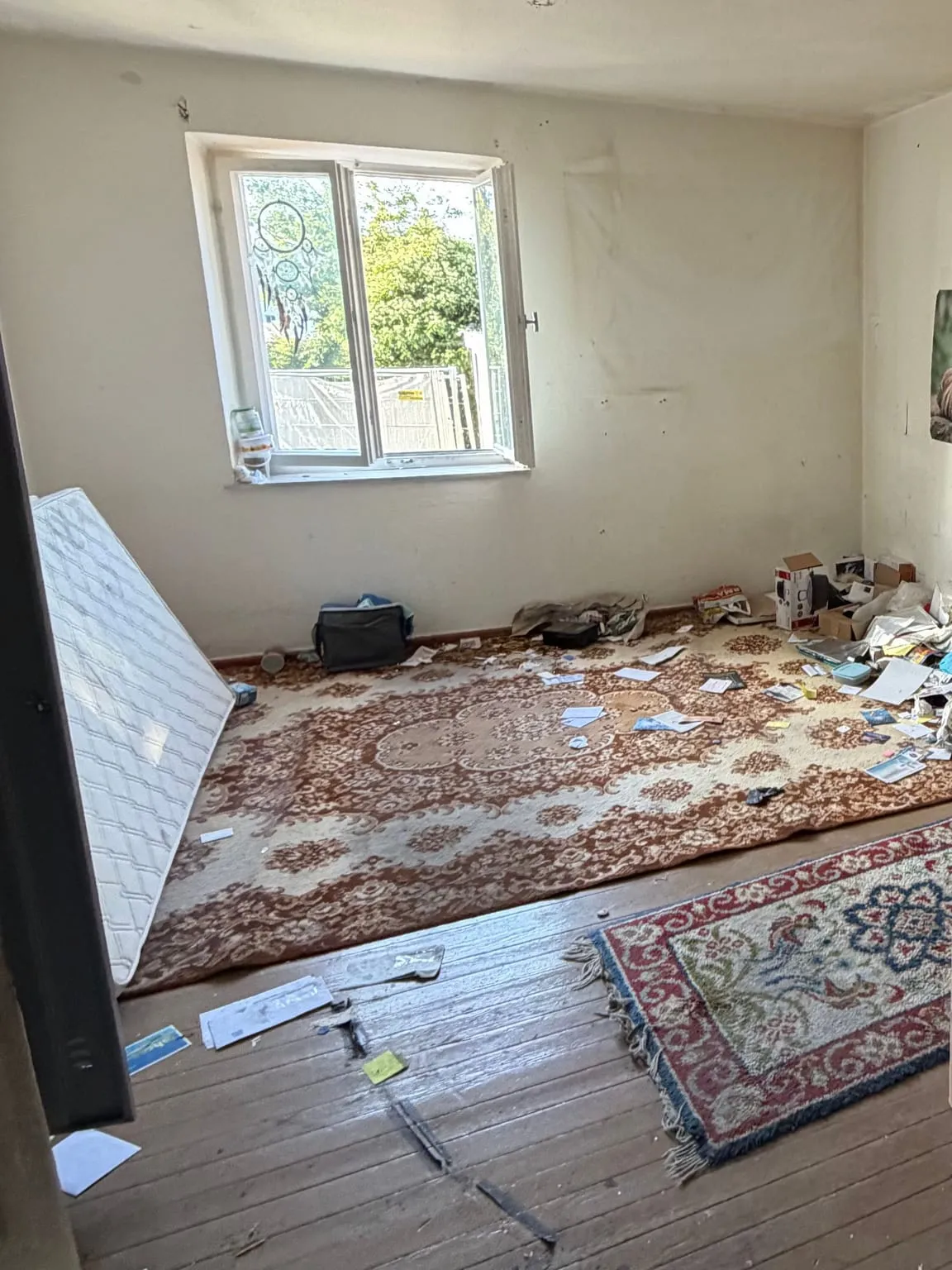 Zimmer mit Fenster an der Stirnwand, auf dem Holzboden liegen zwei Teppiche, eine angelehnte Matratze, Kartons, eine Tasche und verstreute Papiere.
