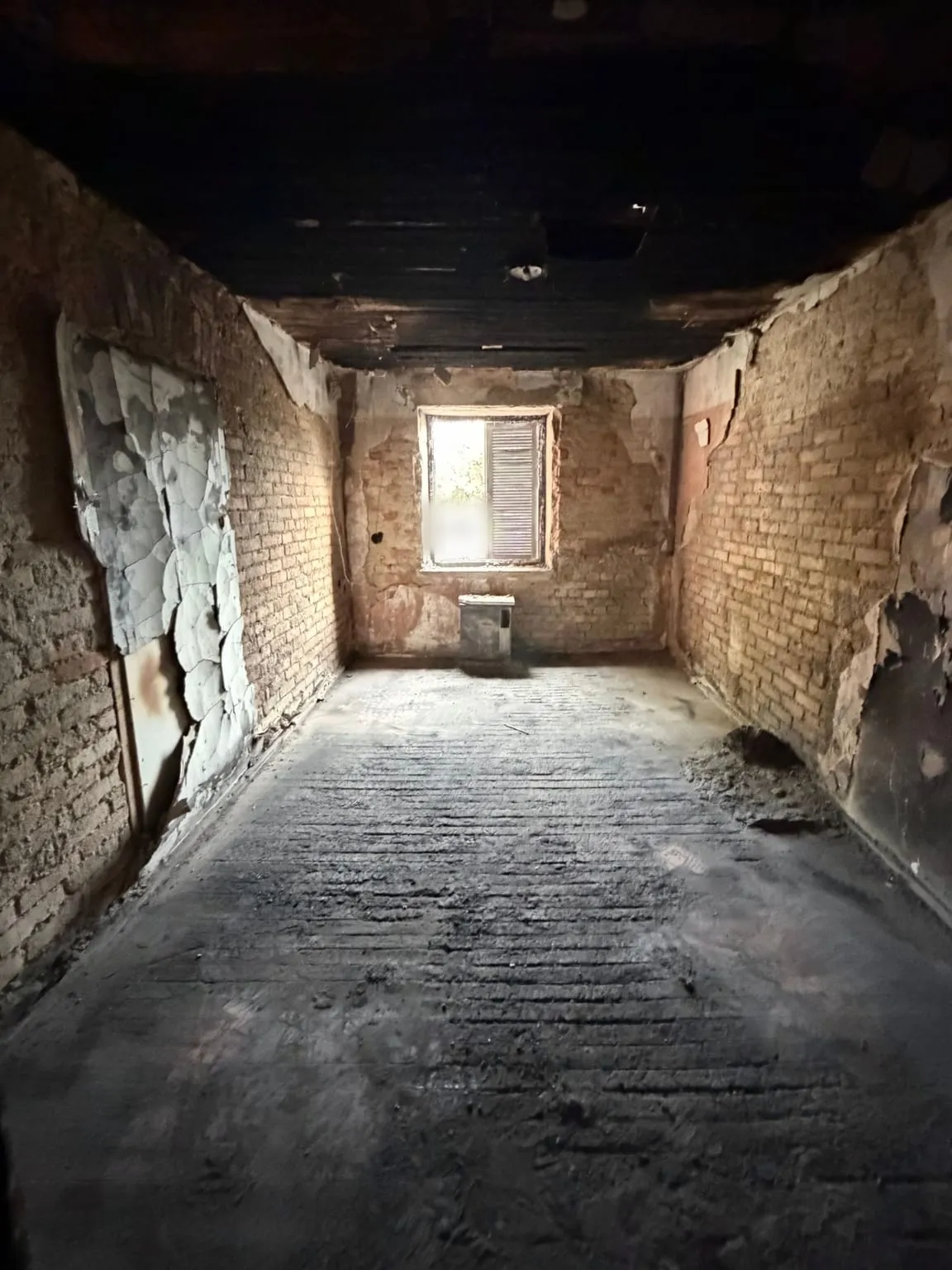 Dasselbe Zimmer mit verkohlter Holzdecke nach der Räumung, der Boden ist frei bis zum Fenster mit Heizgerät an der Rückwand.
