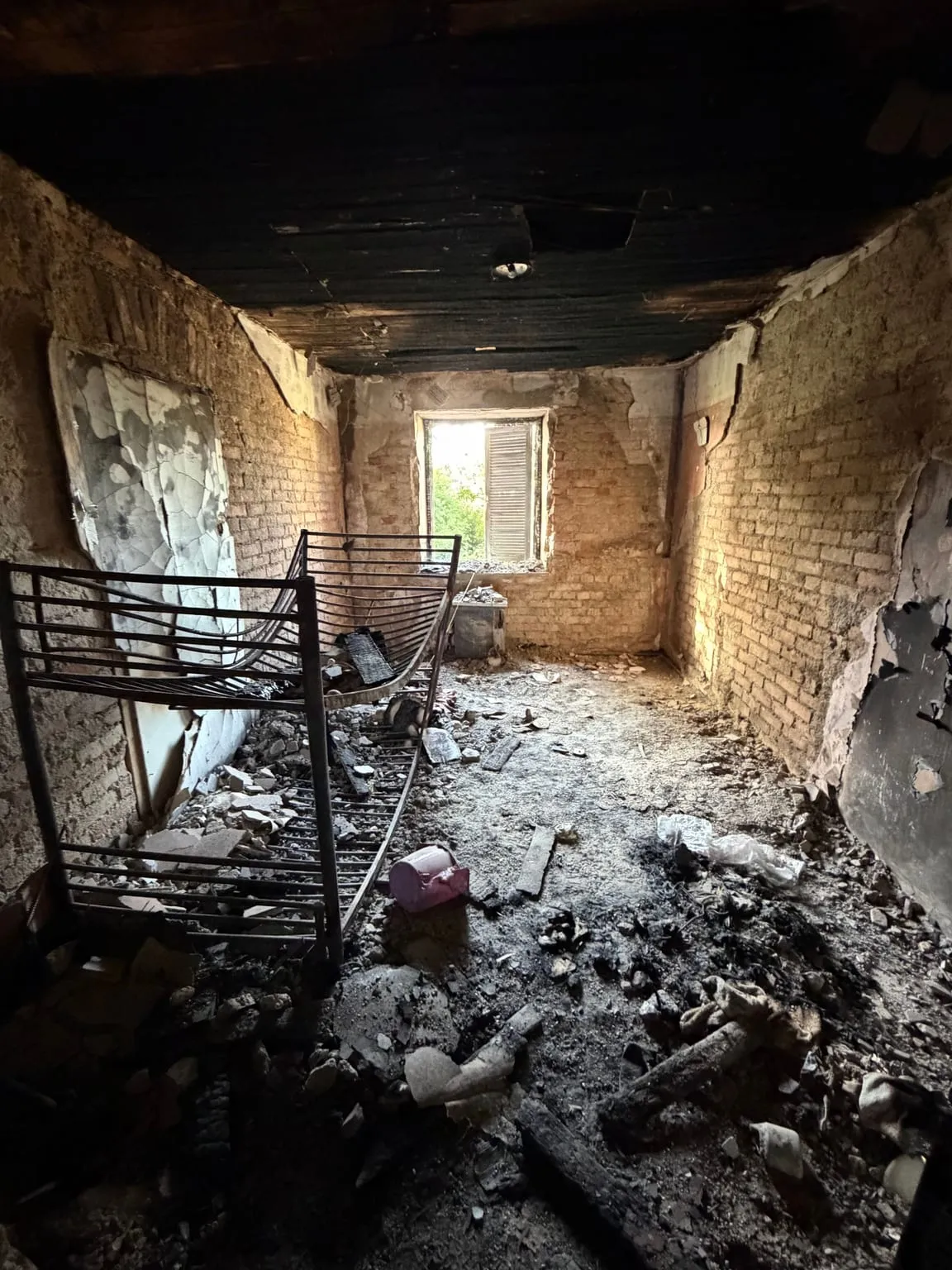 Zimmer mit verkohlter Holzdecke und Ziegelwänden, auf dem Boden ein Metall-Etagenbett, Brandschutt und verkohlte Holzreste.