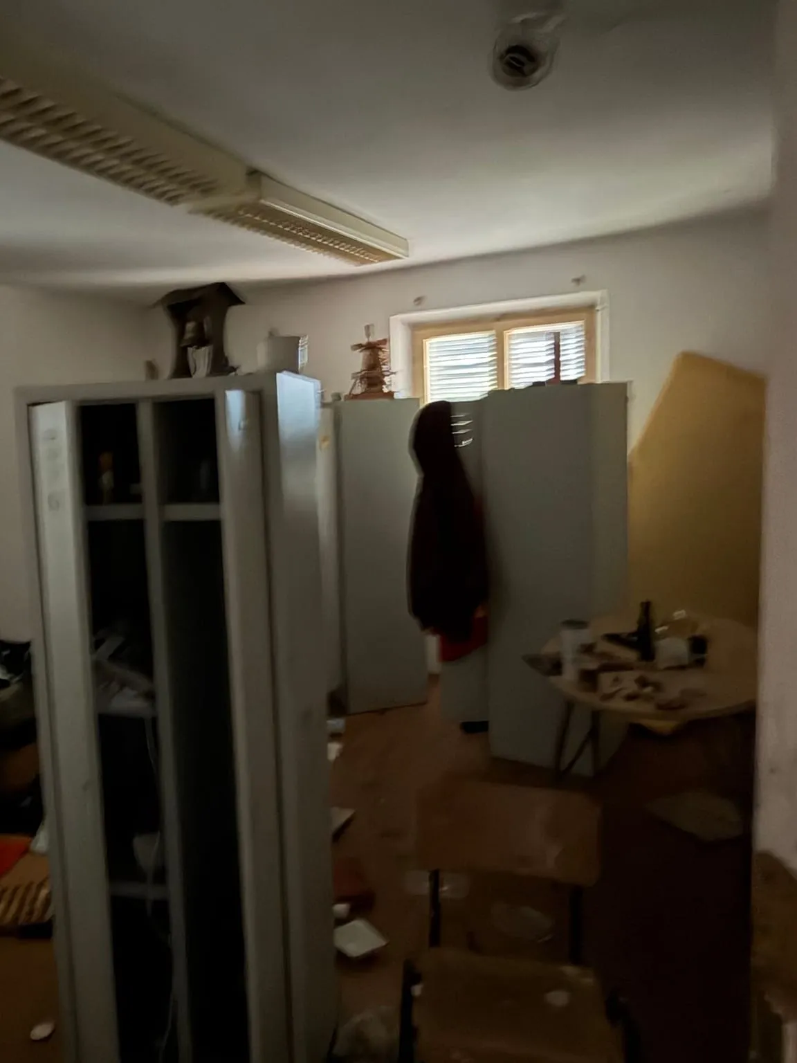 Zimmer mit Deckenleuchte vor der Räumung: Metallspinde, eine Jacke, ein runder Tisch mit Gegenständen und Stühle vor der Fensterwand