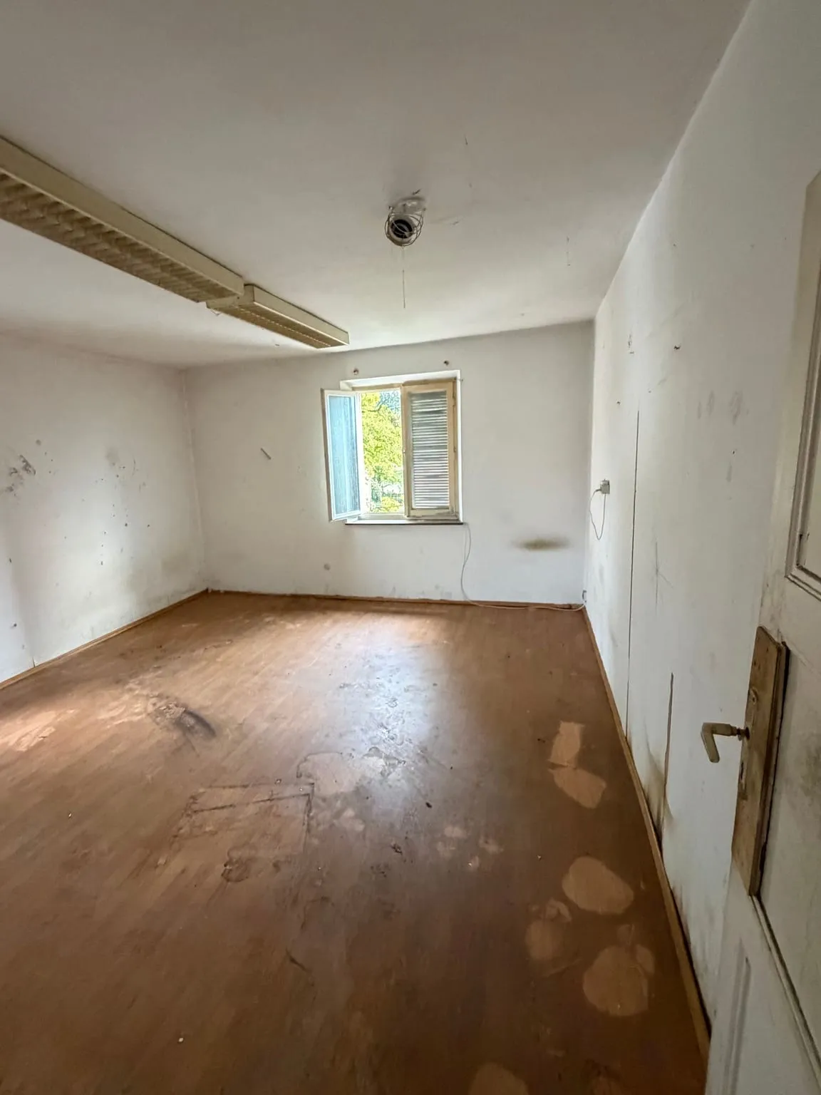 Dasselbe Zimmer leer geräumt, mit freiem Parkettboden, Rasterleuchte an der Decke und geöffnetem Fenster in der Rückwand.