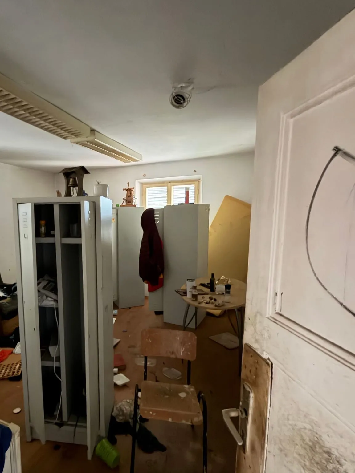 Zimmer mit Rasterleuchte an der Decke, vollgestellt mit Metallspinden, einem Tisch, Stühlen und Gegenständen auf dem Parkettboden, rechts eine geöffnete Tür.