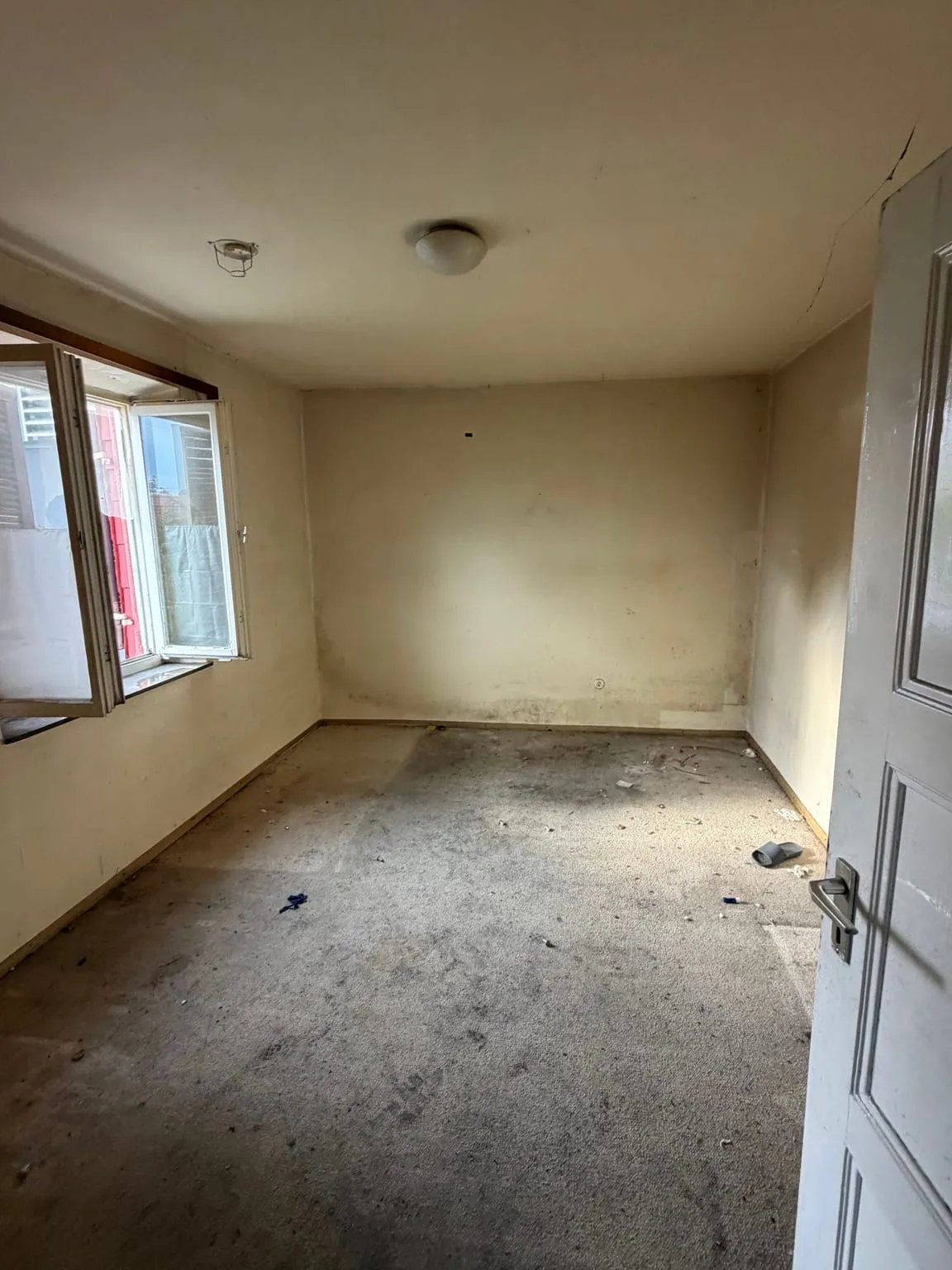 Dasselbe Zimmer nach der Räumung, leer, mit grauem Teppichboden, geöffnetem Fenster links und hellen Wänden.