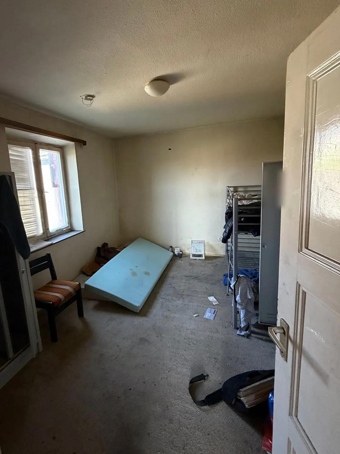 Zimmer mit grauem Teppichboden, links ein Regal und ein Stuhl unter dem Fenster, in der Mitte eine Matratze auf dem Boden, rechts ein Etagenbett aus Metall und ein Spind.