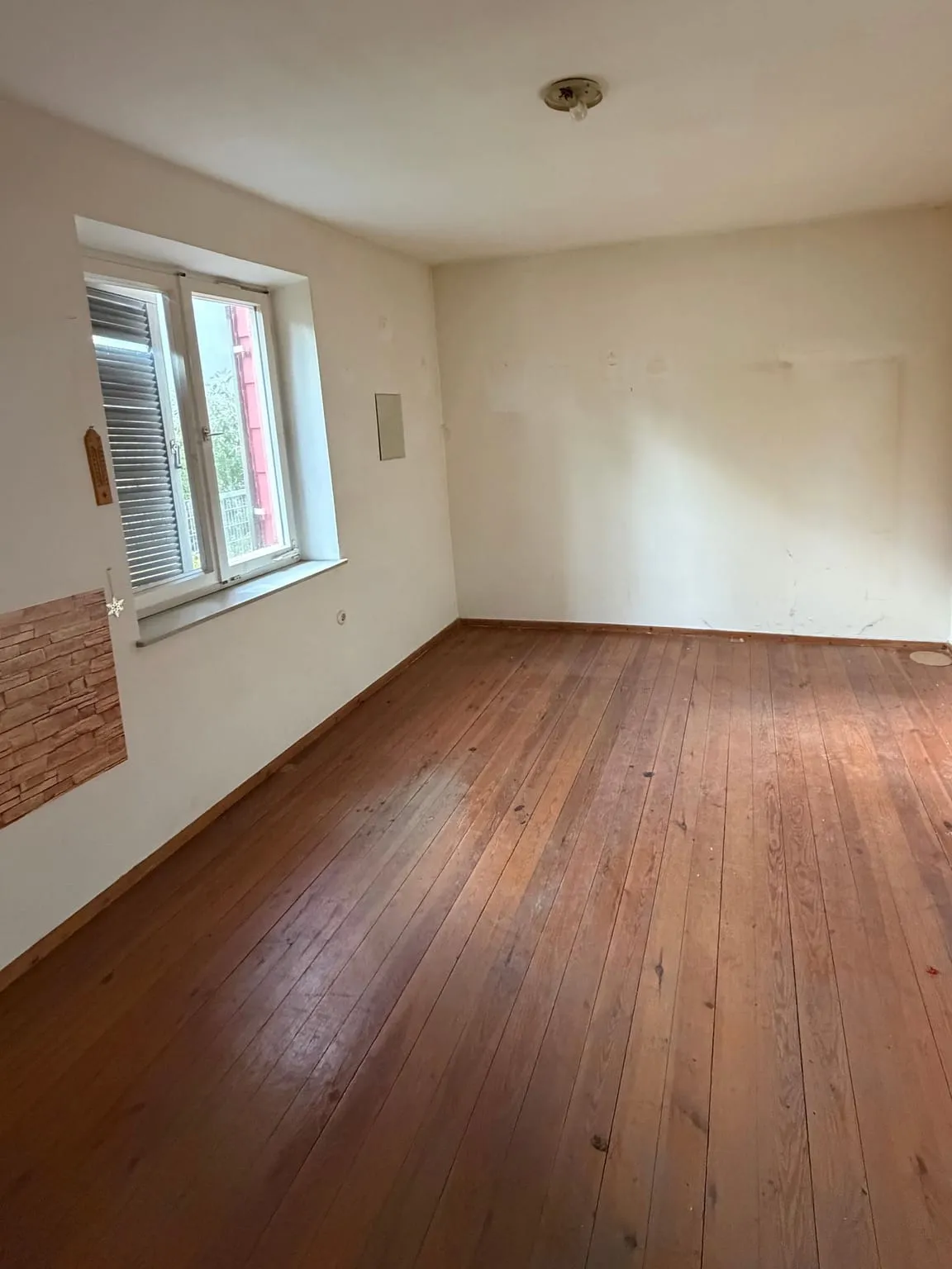 Dasselbe Zimmer leer geräumt, mit freiem Dielenboden, Fenster mit Rollladen und Steinoptik-Paneel an der Wand.