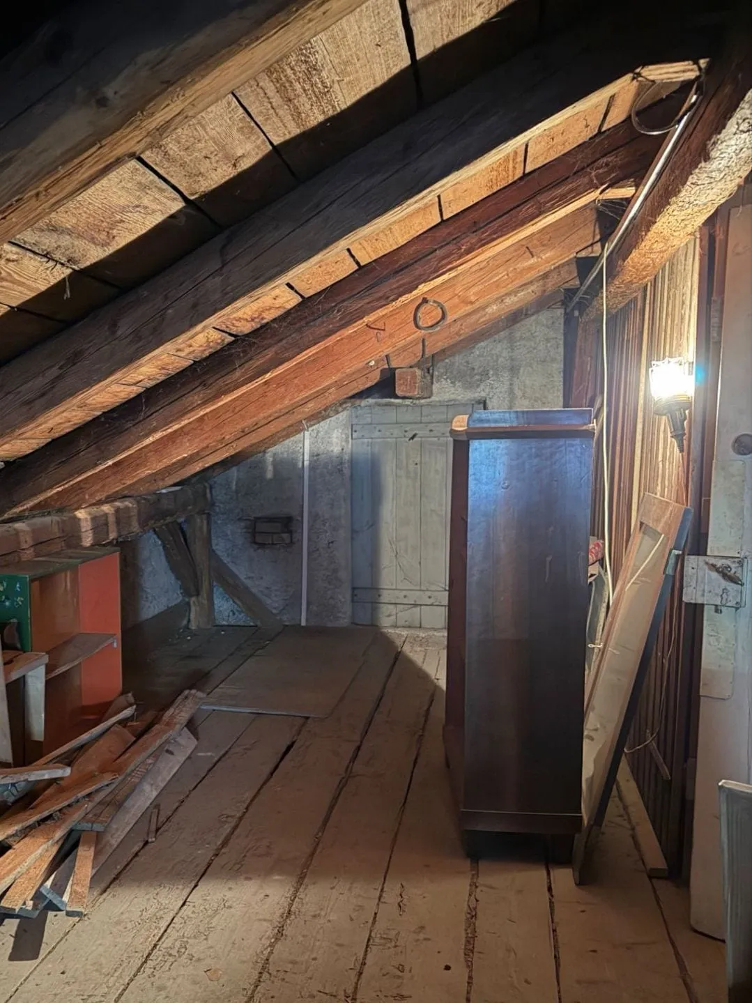 Brettertür in einer gemauerten Wand unter der Dachschräge, rechts davon ein hoher dunkler Kleiderschrank und eine Bretterwand mit Lampe