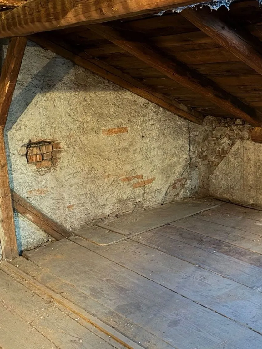 Dieselbe Ecke leer: gemauerte Wand mit kleiner Ziegelöffnung, Holzbalken und freier Dielenboden
