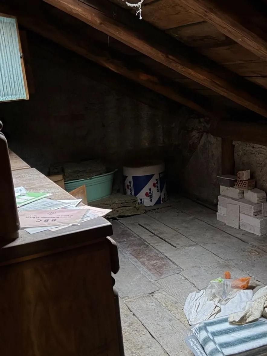 Ecke unter der Dachschräge mit Küchenbuffet, einer Wanne, einer Dämmstoffrolle, Steinen und Farbeimern, davor Tücher und Folien auf dem Boden