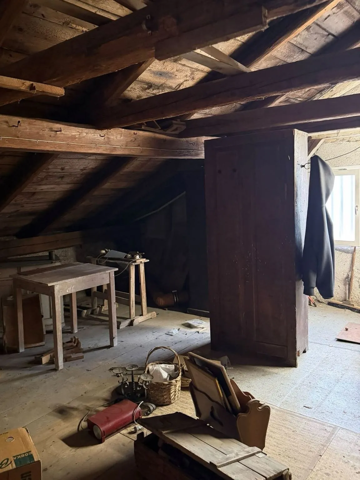 Dachboden mit hohem Holzschrank in der Mitte, dahinter Tisch und Werkbank unter der Schräge, davor Kisten, ein Korb und ein Karton auf dem Boden