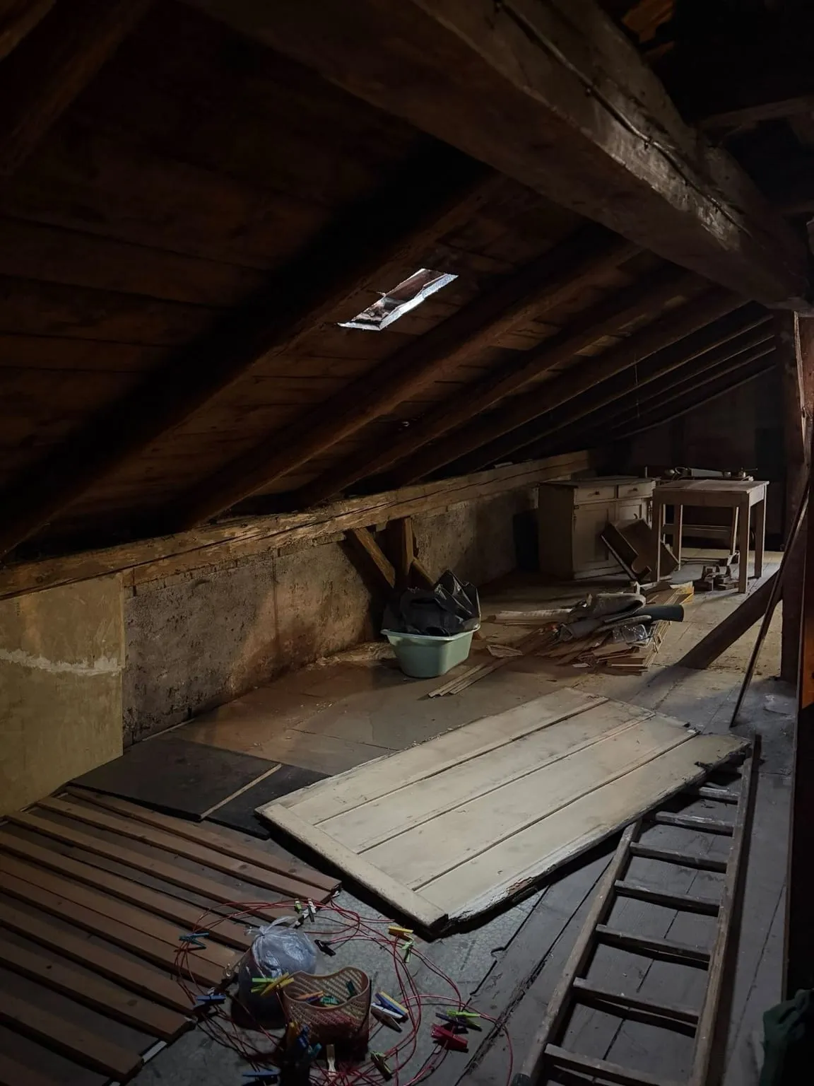 Langer Dachboden unter der Schräge, auf dem Boden ein Lattenrost, eine alte Tür, eine Wäscheleine mit Klammern und eine Leiter, hinten ein weißer Schrank und ein Tisch