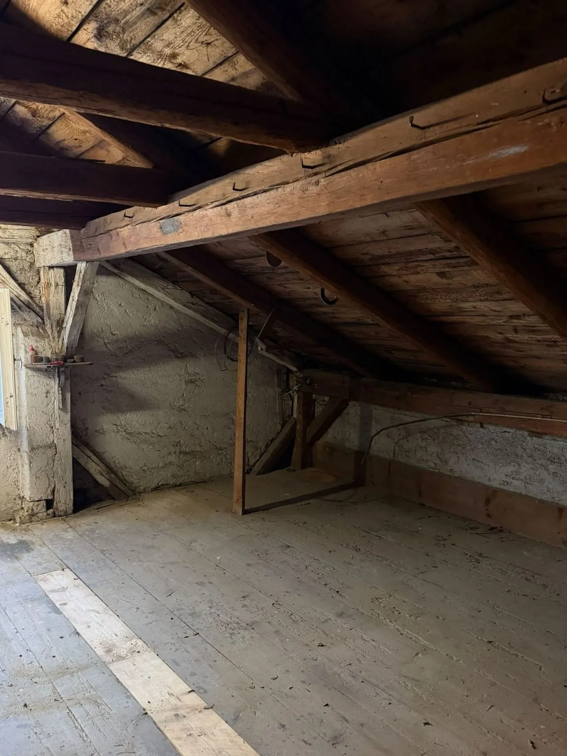 Dieselbe Ecke leer: verputzte Wand mit kleinem Wandbrett, ein dünner Holzpfosten und freier Dielenboden unter dem Dachbalken