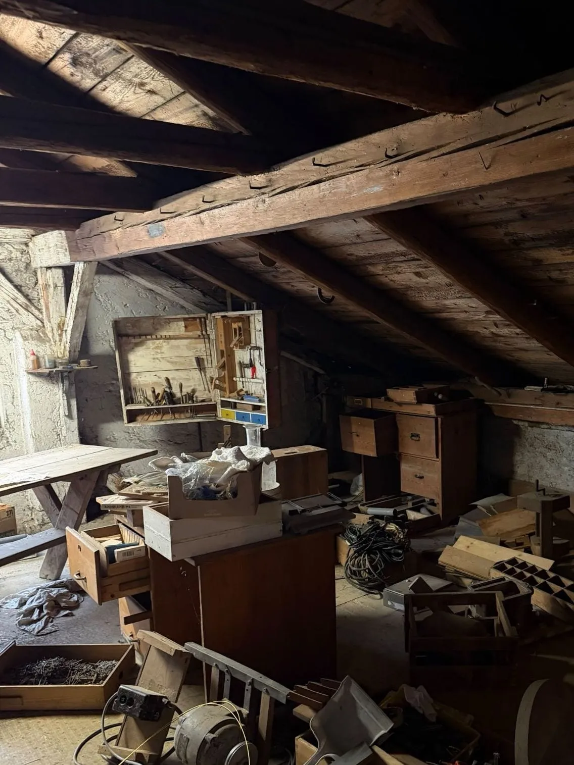 Werkstattecke unter der Dachschräge mit Werkzeugschrank an der Wand, Holztisch, Schubladenkästen, Kabeln, einem Elektromotor und Brettern auf dem Boden
