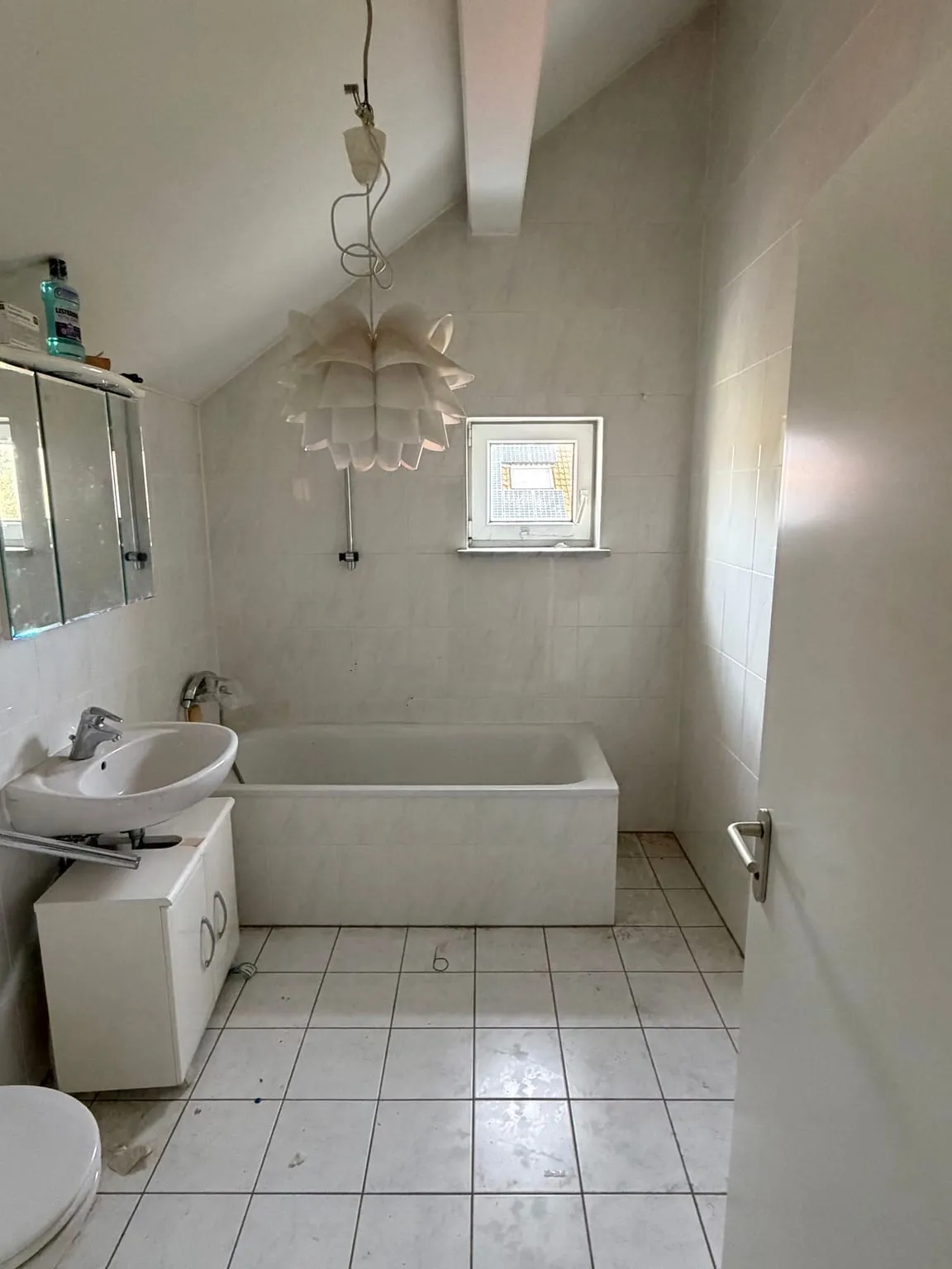 Dasselbe Bad mit Dachschräge nach der Räumung, mit freier Badewanne, Waschbecken, Spiegelschrank und leerem Fliesenboden.