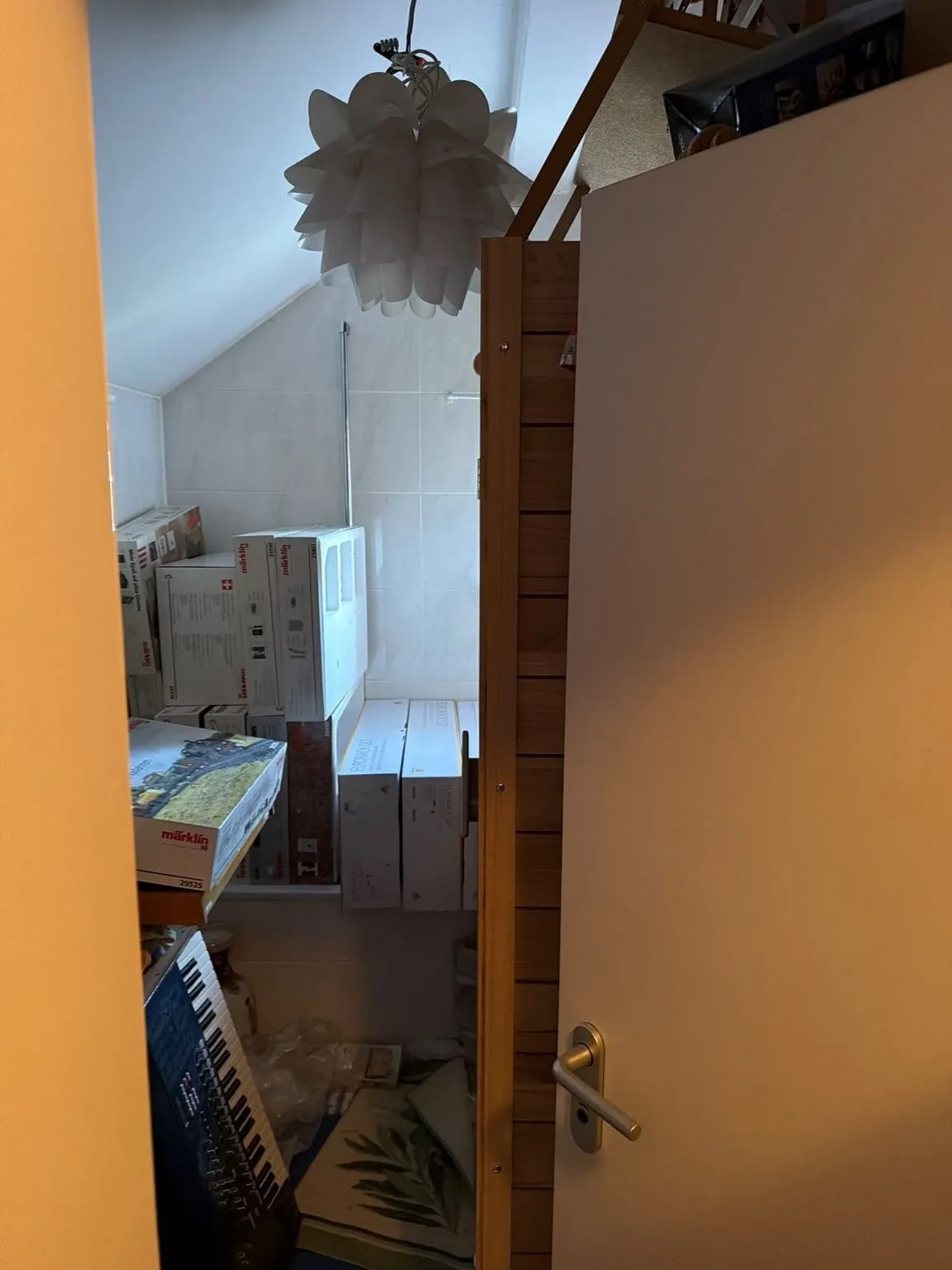 Blick durch die halb geöffnete Tür in ein Bad mit Dachschräge, in dem gestapelte Kartons und ein Keyboard den Bereich vor der Wand verdecken.