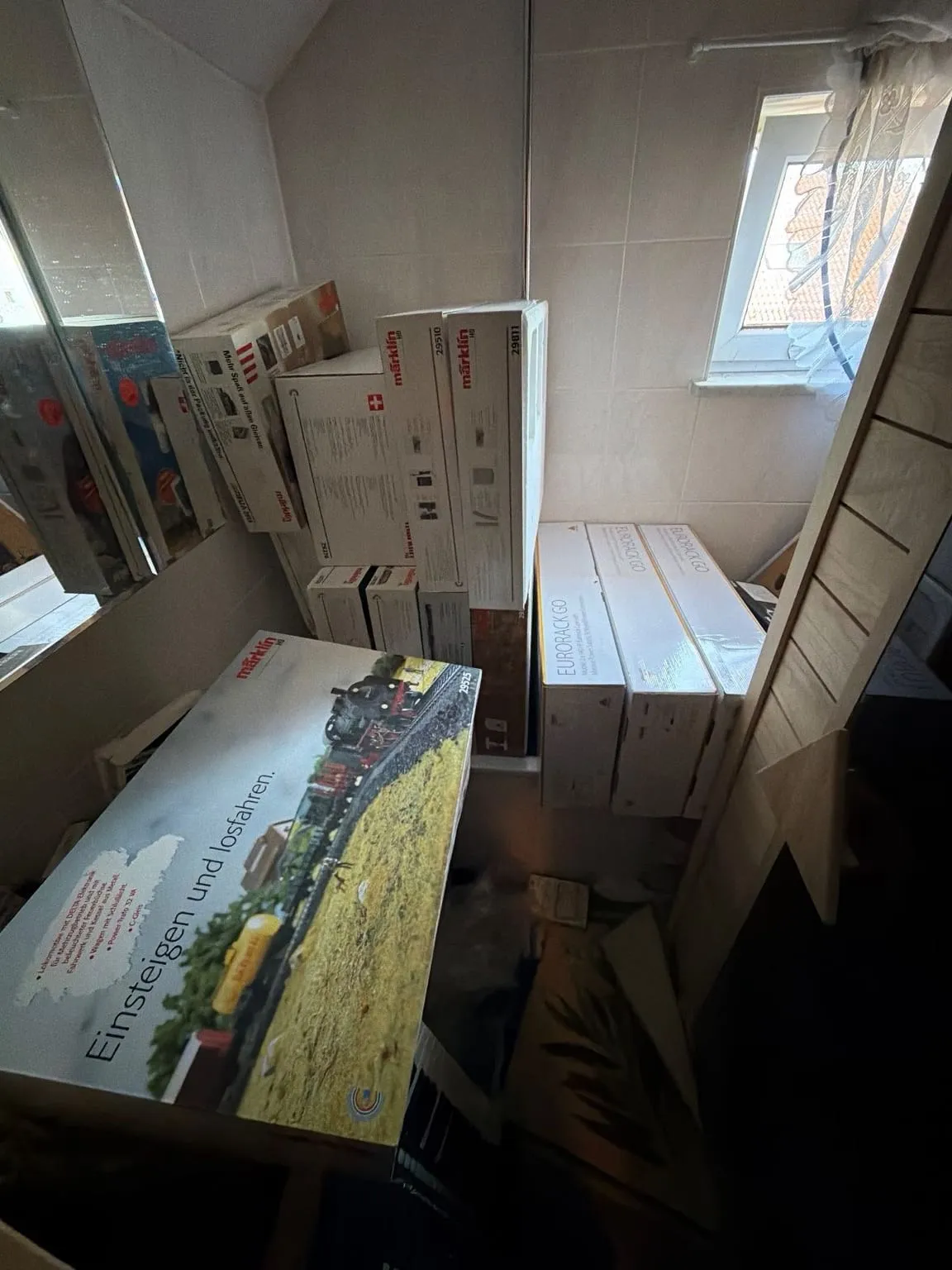 Bad mit Dachschräge vor der Räumung: Kartons stehen gestapelt auf und vor der Badewanne, rechts lehnt ein Holzgestell, am Fenster hängt eine Gardine.