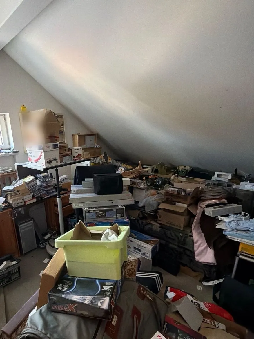 Dachraum vor der Entrümpelung: Unter der Dachschräge stehen Kartons, ein Regal, ein Schreibtisch mit Bildschirmen und Stapeln von Kisten, eine gelbe Kunststoffbox und Modellbau-Kästen.