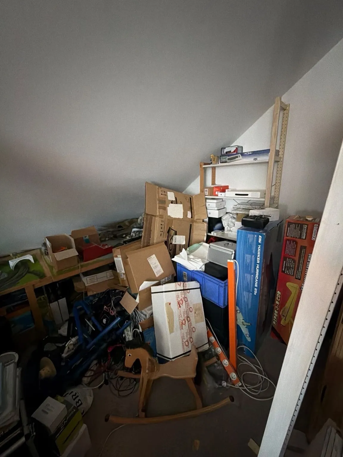 Ecke eines Dachgeschossraums unter der Schräge, zugestellt mit Kartons, einem Holzregal, Modellflugzeug-Kartons, einem Schaukelpferd und Kabeln.