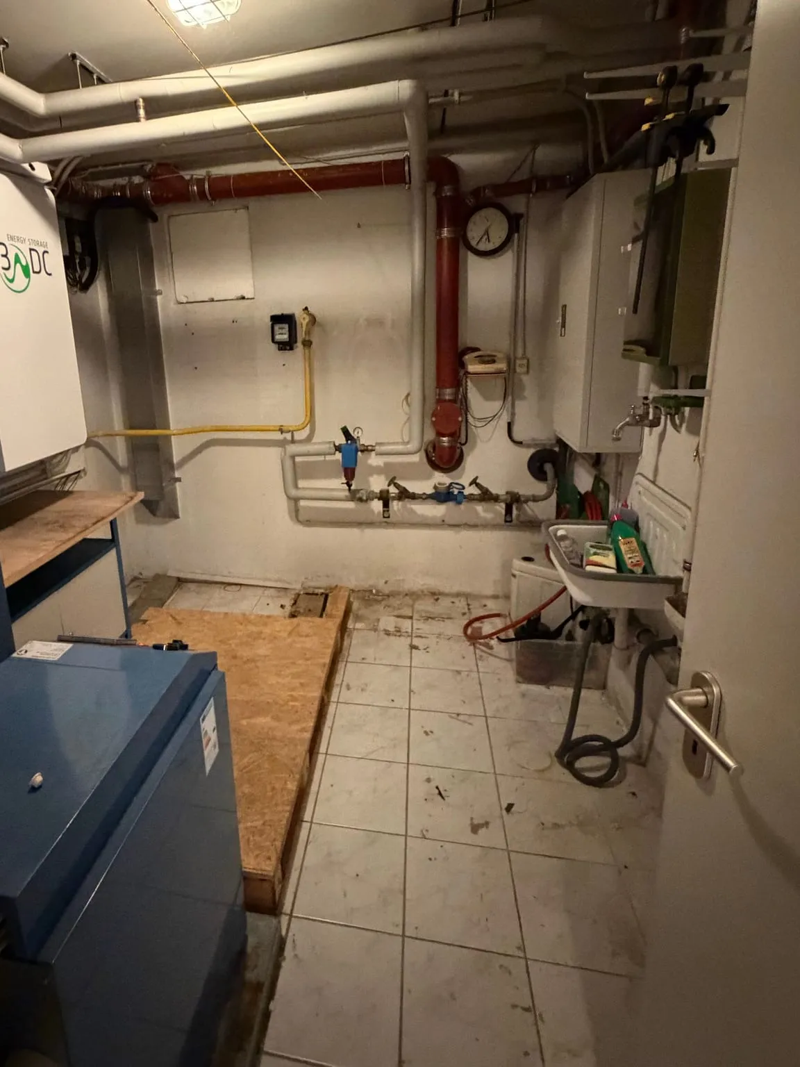 Geräumter Heizungsraum mit blauem Heizkessel, Rohrleitungen an Wand und Decke, Waschbecken und freiem Fliesenboden.