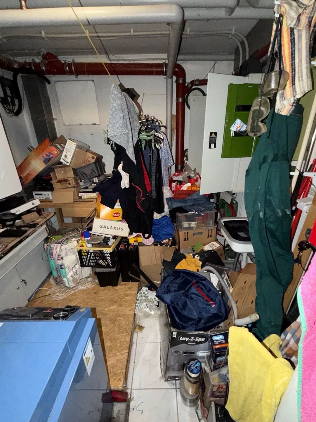 Heizungsraum mit blauem Heizkessel, davor und dahinter Kartons, Kisten, aufgehängte Kleidung und Taschen bis zur Rückwand.
