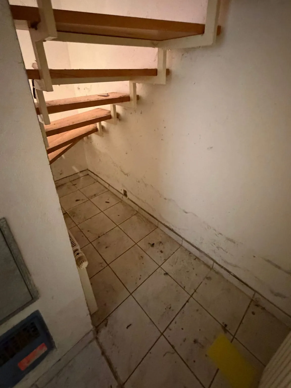 Leere Fläche mit Fliesenboden unter einer offenen Holztreppe, rechts eine weiße Wand, links ein Heizkörper.