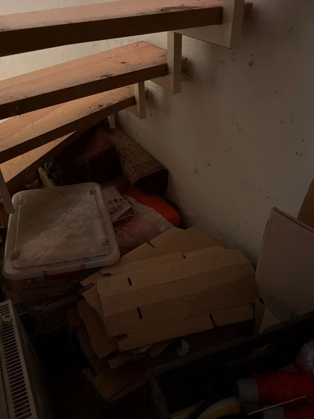Kartons, flach gelegte Pappe, eine Kunststoffbox und Kisten stehen unter einer offenen Holztreppe vor einer weißen Wand.